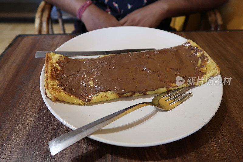 这是一个在白色盘子上涂了一层厚厚的巧克力、两边放着刀叉的卷好的甜早餐煎饼/可丽饼的形象